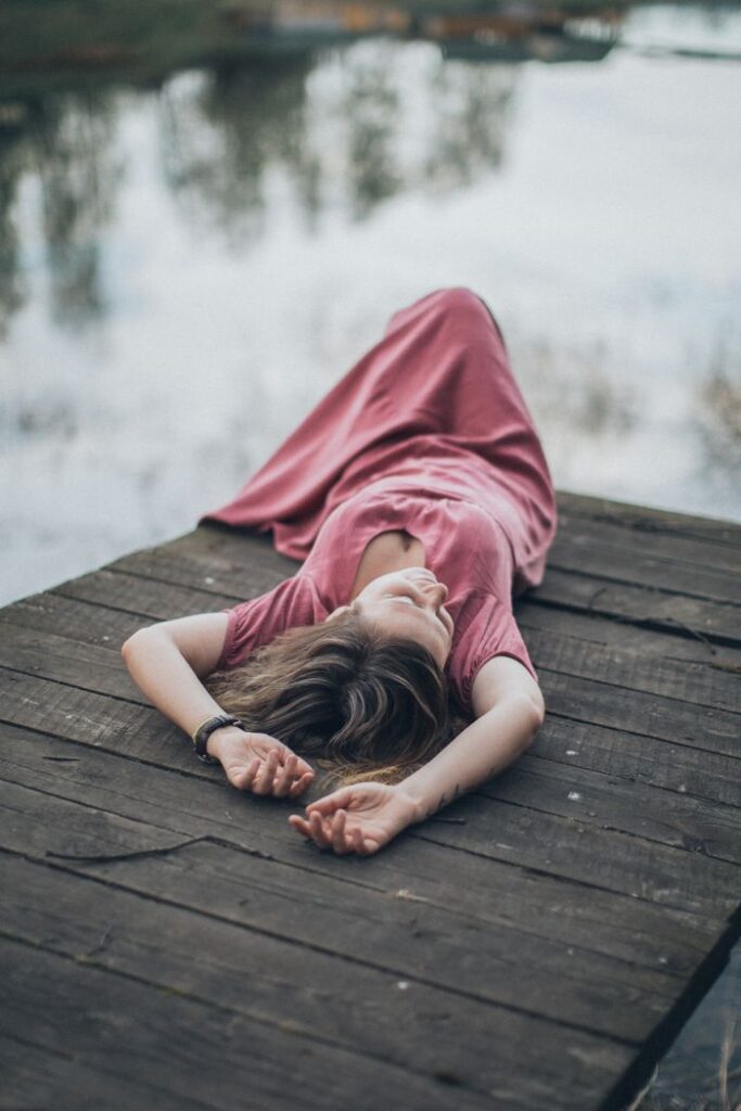Woman in a pink dress lying on a wooden dock by the water, enjoying a calm moment outdoors.